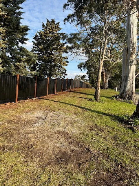 Long black Colorbond fence with wooden posts running alongside trees and a grassy area