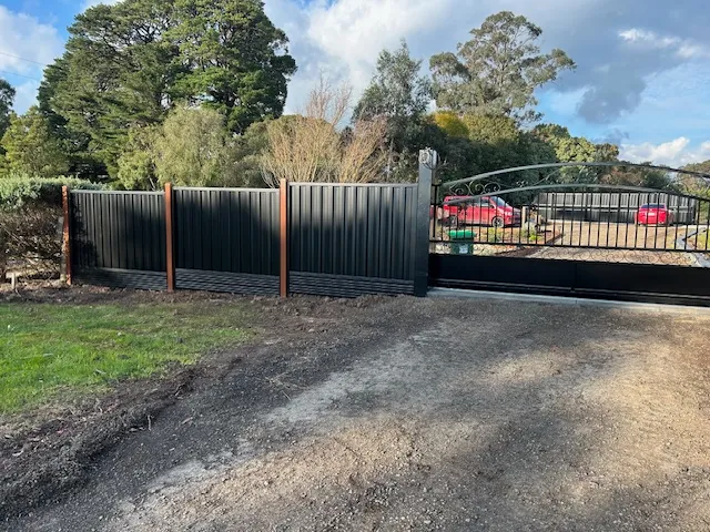 Black Colorbond fence with wooden posts beside a black metal driveway gate leading to a house and garden