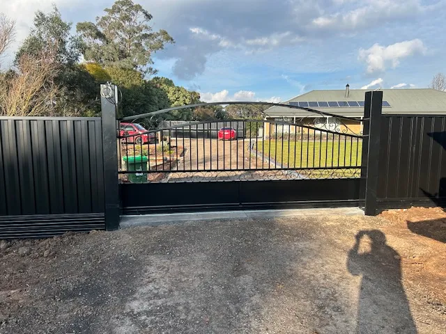Black Colorbond fence with matching metal driveway gate leading to a house with solar panels and gravel path